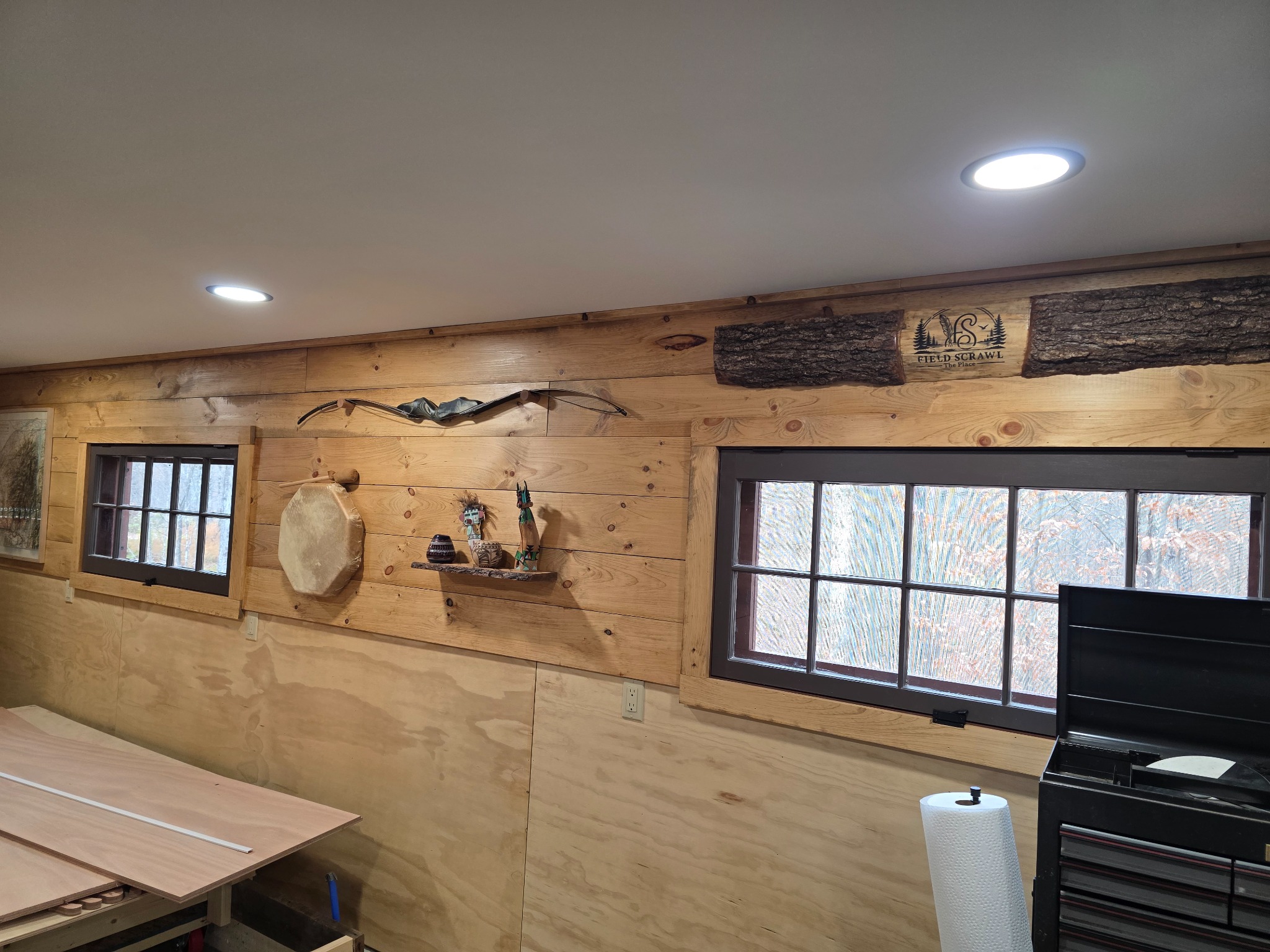Completed workshop wall with a bow above a rawhide drum, pottery and carvings on a maple shelf; boatbuilding boards below